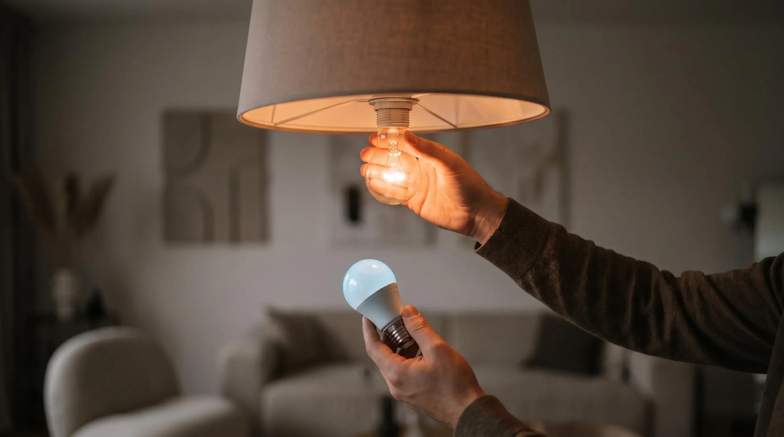 Main remplaçant une ampoule halogène par une ampoule LED dans un salon lumineux, symbolisant la transition énergétique du foyer